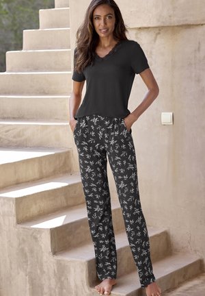 Schwarzes kurzärmliges Oberteil mit Spitzenhalsausschnitt, kombiniert mit schwarzen Hosen mit einem weißen Blumenmuster und seitlichen Taschen, die auf einer Treppe stehen.