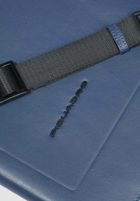 Bolso de cuero azul texturizado con correa negra ajustable y logo "PIQUADRO" negro en relieve centrado en la superficie.