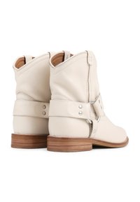 Botas de tobillo de cuero crema con punta redondeada, caña corta y tacón de madera. Presenta una correa envolvente con acentos metálicos.