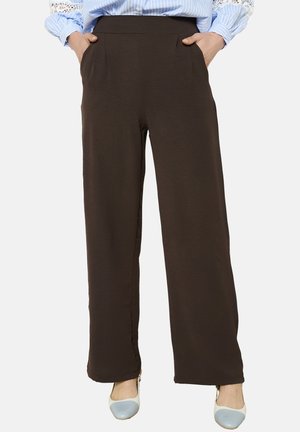 Pantaloni a gamba larga marroni con una texture liscia, caratterizzati da una vita alta e tasche laterali. Indossati con calzature accentate di blu chiaro.