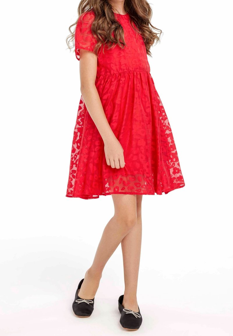 Robe rouge à manches courtes, taille froncée, avec un voile transparent présentant un motif floral subtil. Associée à des chaussures noires ornées de nœuds.