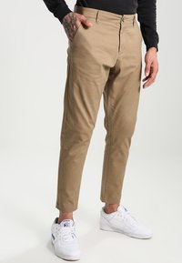 Beżowe chinosy o prostym kroju, z suwakiem i szlufkami na pasek, zestawione z białymi trampkami, przedstawione na neutralnym tle.