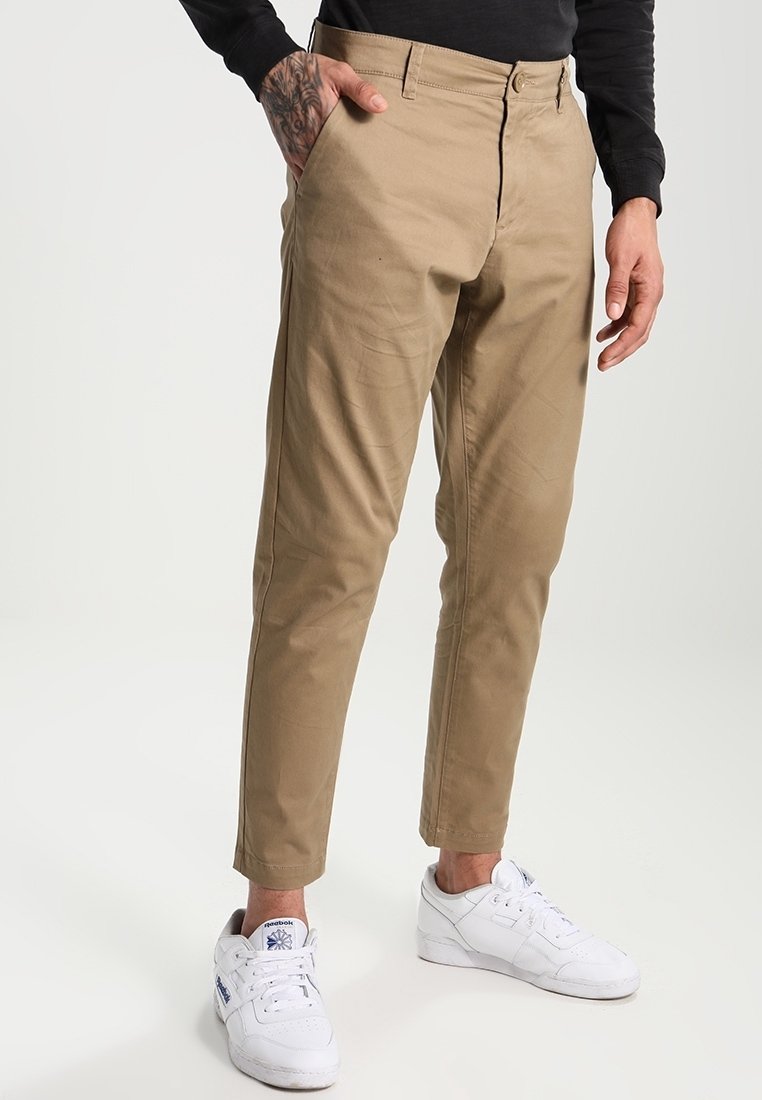 Beżowe chinosy o prostym kroju, z suwakiem i szlufkami na pasek, zestawione z białymi trampkami, przedstawione na neutralnym tle.