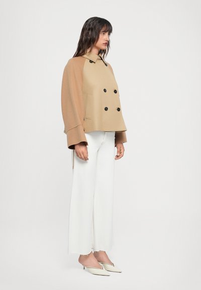 Beige double-breasted jacket with oversized sleeves, black buttons, and side pockets, paired with white flared jeans and off-white heels.