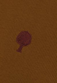 Tejido marrón texturizado con un pequeño logotipo de árbol bordado en color marrón oscuro. La superficie está tejida con un ligero estiramiento, ofreciendo durabilidad y flexibilidad.