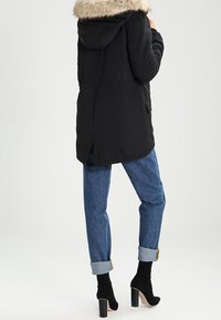Czarna parka z futrzanym kapturem, z zaznaczoną talią, bocznymi kieszeniami i dłuższym tylnym hemem, zestawiona z jeansami w niebieskim kolorze z podwiniętymi mankietami.