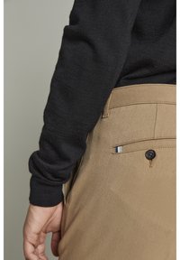 Czarny rękaw swetra obok beżowych spodni z zapięciem na guzik na tylnej kieszeni, z małym akcentem w paski. Materiał wygląda na gładki i dobrze ułożony.