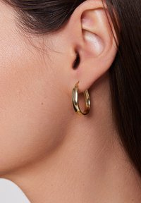 Złota kolczyk w kształcie kółka zaprezentowany na uchu modelki, o gładkiej, błyszczącej powierzchni i okrągłym kształcie. Średnica jest średniej wielkości.