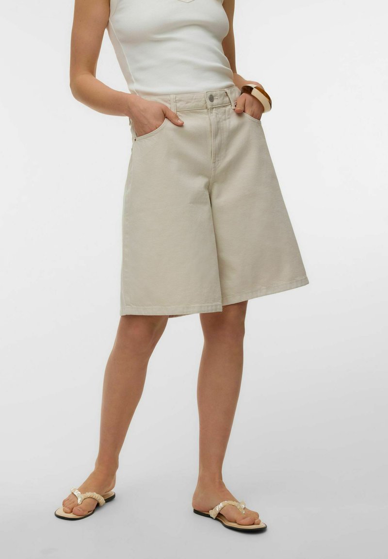 Femme portant un short large beige jusqu’aux genoux, un débardeur blanc, des sandales beige, les mains dans les poches sur fond blanc.