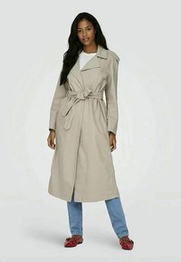 Casaco trench bege com cinto na cintura, gola tipo notch e mangas compridas, combinado com jeans azul claro e sapatos vermelhos de fácil calçar.