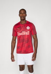 Maillot de soccer rouge à manches courtes avec un motif graphique, col rond et accents de logo. Assorti à un short blanc, mettant en valeur un design sportif.