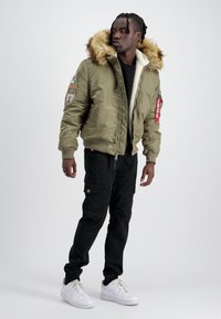 Giubbotto bomber verde oliva con cappuccio in pelliccia sintetica, polsi a costine e toppe. Abbinato a pantaloni cargo neri e sneakers bianche.