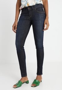 Jeans de mezclilla de corte ajustado en azul oscuro con costuras en contraste, cintura alta, cinco bolsillos y una textura suave. Combinados con mules de charol verdes.
