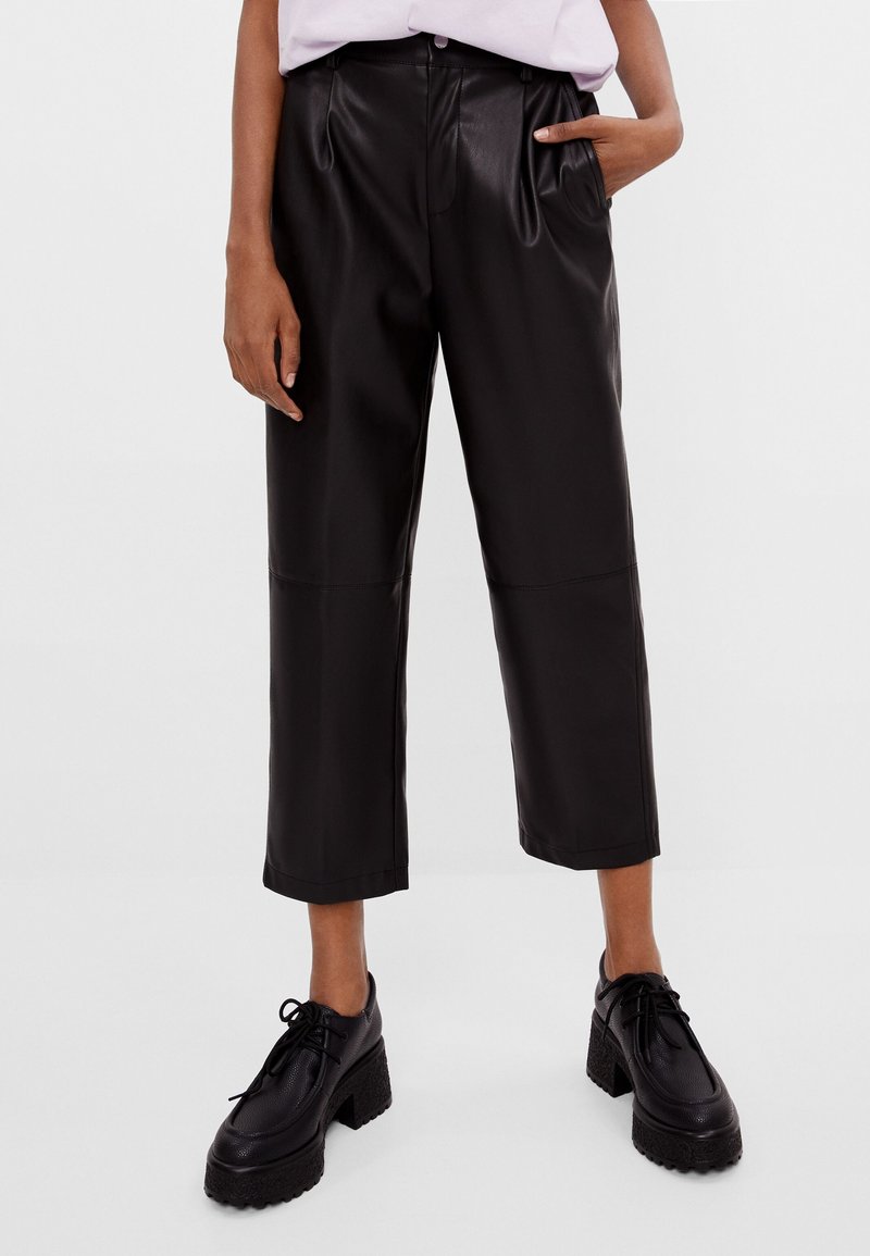 Personne portant un pantalon court taille haute en faux cuir noir avec des plis, associé à des chaussures à plateforme noires à lacets épaisses.