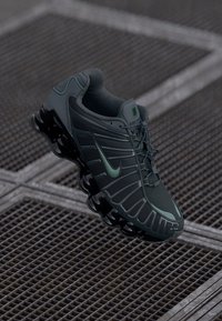 Tamnozeleni i crni Nike tenisice za trčanje s jedinstvenom segmentiranom potplatom, prikazane na pozadini od metalnih rešetki.