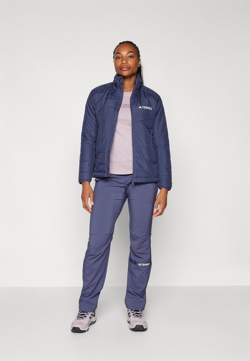 Doudoune navy avec col montant et logo, associée à un t-shirt violet clair et un pantalon navy assorti. La chaussure est grise avec des accents noirs.