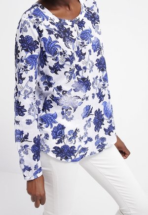 Personne noire portant un haut blanc à manches longues avec des motifs floraux bleus et un pantalon blanc ajusté, raccourci à mi-cuisse.