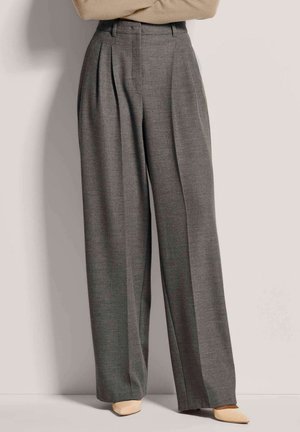 Grijze broek met hoge taille en wijde pijpen met plooien, gecombineerd met beige puntige schoenen, gedragen door een persoon met gekruiste armen.