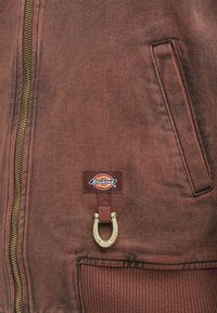 Καφέ βαμβακερό μπουφάν με χρυσό φερμουάρ, μπροστινή τσέπη και λογότυπο με σχέδιο πέταλου στην αριστερή πλευρά. Υφή υφάσματος.