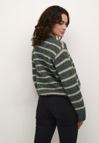 Pulover tricotat verde și bej cu dungi, cu un croi lejer, gât rotund, mâneci lungi și manșete răsucite, asortat cu blugi închiși la culoare.