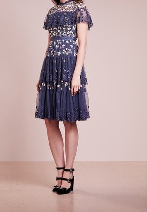 Vrouw draagt een marineblauwe, knielange jurk met geplooide lagen en bloemrijke borduursels, gecombineerd met zwarte pumps met enkelbandjes.