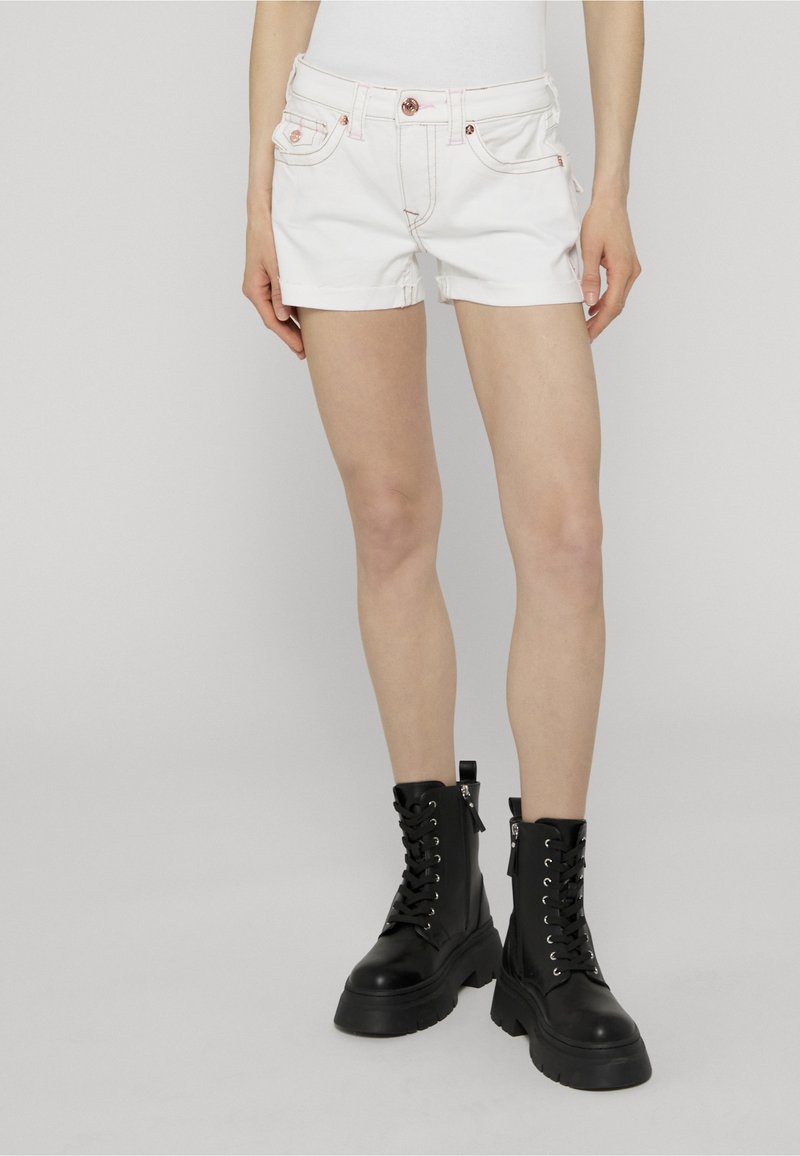 Personne portant un short en jean blanc et des bottes de combat noires à lacets, debout devant un fond neutre.