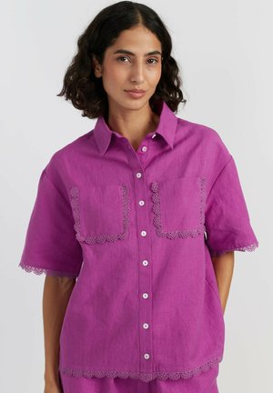 Femme portant une chemise boutonnée violet vif à manches courtes avec bordure en dentelle sur les poches, les manches et l'ourlet, sur un fond uni.