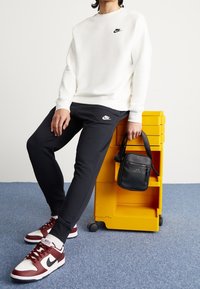 Biały sweatshirt Nike, czarne spodnie dresowe Nike, czerwone i białe sneakersy Nike, czarna torba na ramię z zamkiem, żółta wózek do przechowywania.