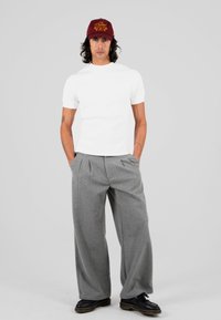 Młody mężczyzna z długimi, kręconymi włosami, noszący czerwoną czapkę, białą koszulę z krótkim rękawem, szare szerokie spodnie i czarne buty, stoi na szarym tle.