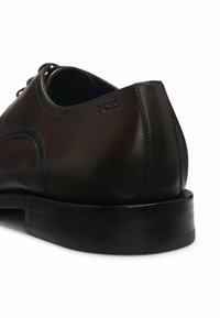 Scarpa elegante in pelle marrone con texture liscia, punta arrotondata, design con lacci e discreto logo sul tallone. Suola robusta in gomma nera.