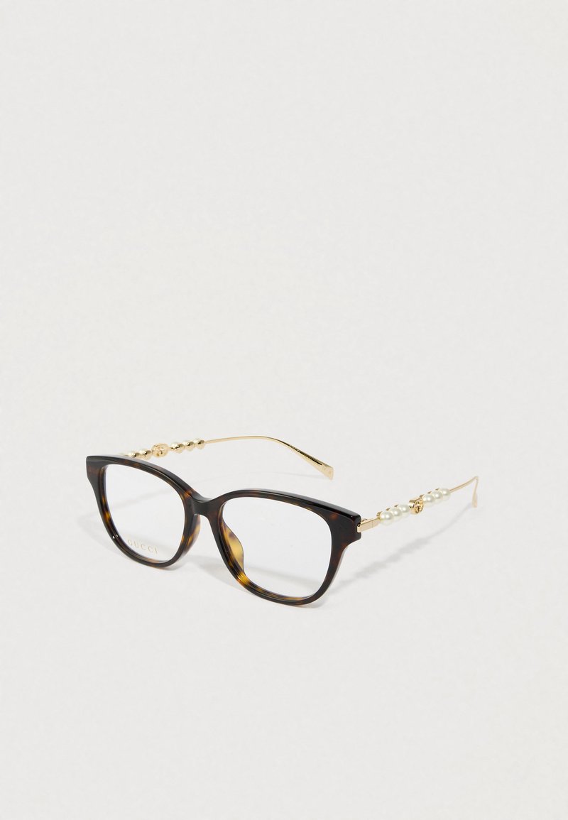 Óculos de tartaruga com armações quadradas, hastes douradas decoradas com pérolas e pequenos ornamentos dourados sobre um fundo branco.