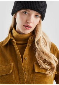 Jongedame met lang blond haar, die een zwarte gebreide muts, een mosterdkleurige coltrui en een corduroy jas met voorzakken draagt, kijkt lichtjes naar voren.
