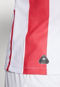 Rød og hvit sportsdrakt med chevronmønster, med en svart "Kombat System" logoetikett nederst til venstre.