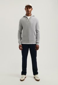 Lichtgrijze gebreide pullover met een halve rits kraag, gecombineerd met marineblauwe broek en witte sneakers. Heeft een ribbelstructuur en lange mouwen.
