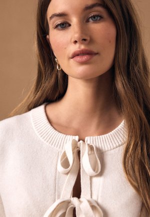 Mladá žena s dlhými hnedými vlasmi, ktorá nosí zlaté kruhové náušnice a krémový pletený sveter previazaný dvoma mašľami na výstrihu.