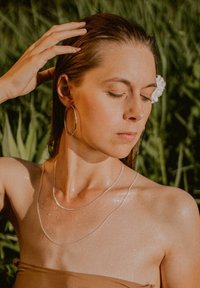 Silver layered necklaces and large hoop earrings adorn a person with wet skin, set against green foliage in soft daylight.
