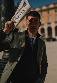 Žalias švarkas per juodą megztinį, baltą marškinių apykaklę ir pilką kaklaraištį. Laiko laikraštį virš galvos prieš saulėtą miesto foną.