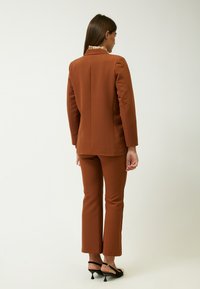 Blazer marrone su misura e pantaloni slim-fit coordinati, realizzati in tessuto morbido; scarpe nere con dettagli decorativi completano l'insieme.