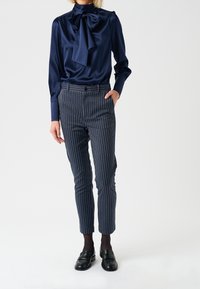 Blusa in satin blu marina con fiocco, abbinata a pantaloni blu con righe sottili. I pantaloni presentano una vestibilità sartoriale con orlo a risvolto. Mocassini neri completano il look.