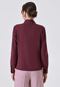 Bordeauxrote Bluse mit langen Ärmeln und einem Kragen, mit einem Knopfdetail auf der Rückseite. Glattes Material, ergänzt durch hellrosa Hosen.