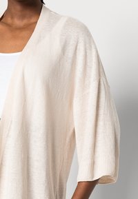 Cardigan beige clair en maille ouverte avec des manches courtes, doté d'un poignet côtelé et d'une texture douce. Porté sur un haut blanc.