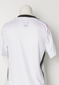 Bijela Adidas sportska majica s crnim ovratnikom, kratkim rukavima i mrežastim bočnim panelima, s oznakom veličine S na stražnjoj strani.