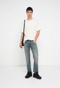 Μπεζ ριμπ μπλούζα με κοντά μανίκια, ανοιχτό μπλε τζιν και μαύρα sneakers. Το μοντέλο κρατά μια μαύρη τσάντα ώμου, στέκεται μπροστά σε λευκό φόντο.