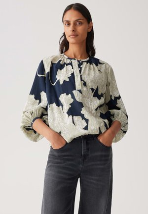 Woman wearing a loose floral blouse with puffed sleeves and black jeans, hands in pockets, standing against a plain light background.