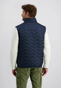 Marineblauwe gewatteerde vest met een hoge kraag, voorzien van een zigzagpatroon. Gedragen over een gestructureerd witte trui, gecombineerd met groene corduroy broek.