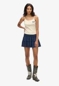 Top de tirantes color beige con detalles de encaje y botones, combinado con una falda azul marino de volantes; la modelo lleva botas de cowboy grises con adornos y estrellas blancas.