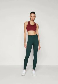 Reggiseno sportivo color bordeaux, abbinato a leggings verdi a vita alta. I leggings sono aderenti con una texture liscia; entrambi i capi sono funzionali ed elasticizzati.