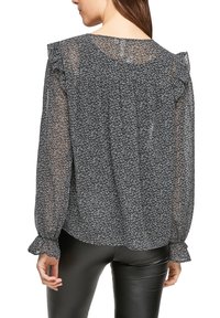 Blouse noire transparente avec un motif floral blanc, détails froncés aux épaules et poignets élastiques. Portée avec un pantalon noir ajusté.