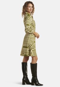 Groene jurk met een patroon van geometrische vormen en lijnen, lange mouwen en een uitlopende rok, gecombineerd met kniehoge zwarte laarzen.