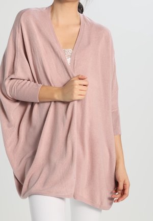 Femme portant un cardigan rose pâle, ample et doux, par-dessus un top en dentelle blanche et un pantalon blanc, tenant le devant du cardigan d'une main.